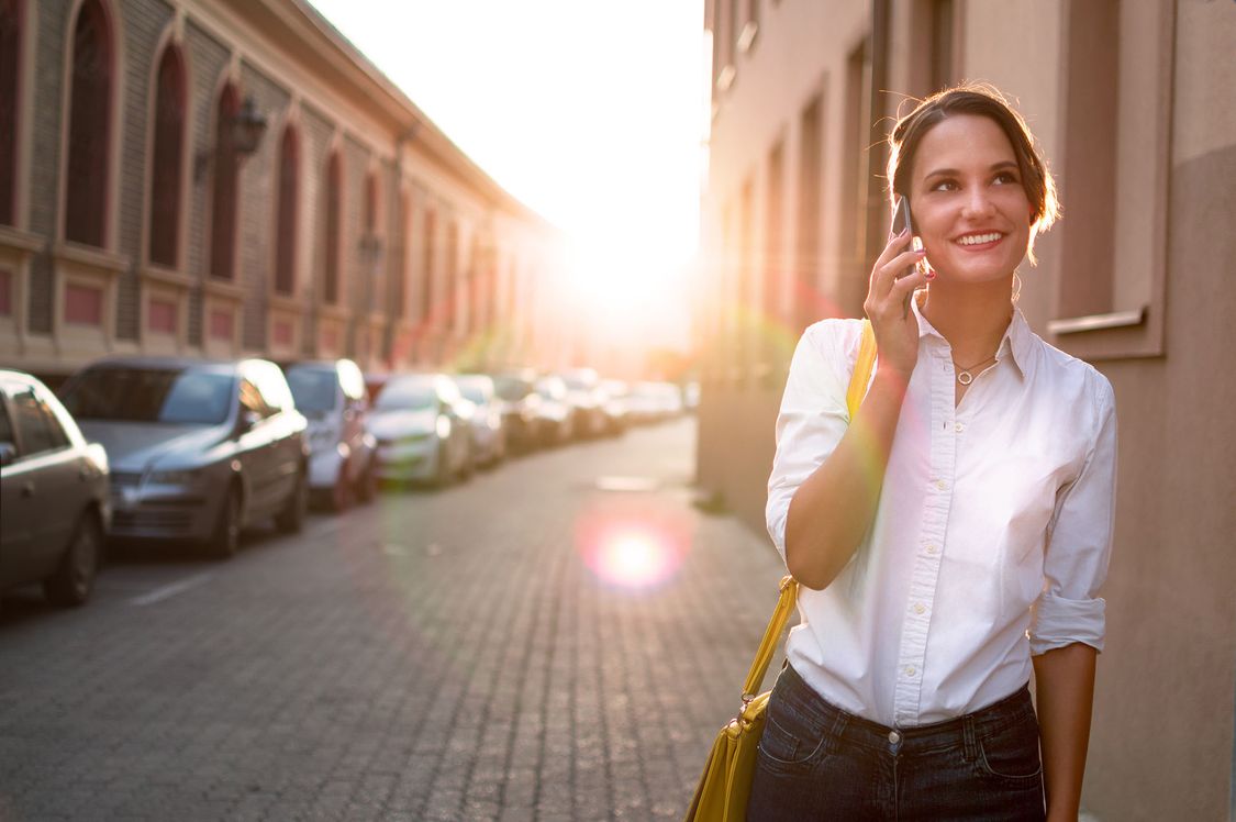 Eine junge Frau steht neben einer Straße mit urbanem Charakter und telefoniert mit ihrem Smartphone