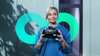 Woman in blue shirt holding VR headset, teal logo behind vr-headset