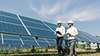 two employees wearing hard hats are walking near solar panels