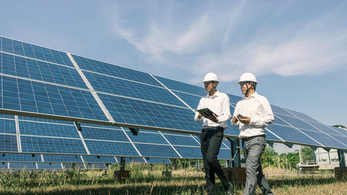 two employees wearing hard hats are walking near solar panels 