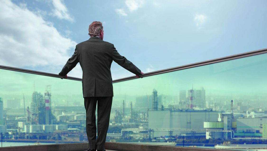 We see the back of a man standing at a transparent parapet, high above a city that he’s looking down on.