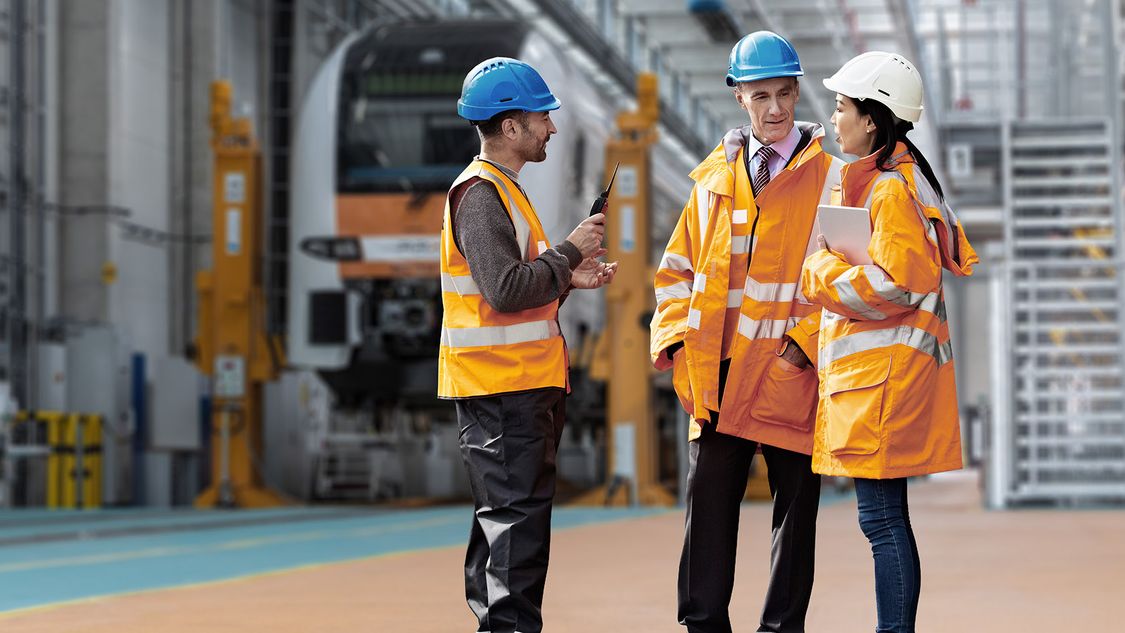 Three representatives of the Alliance for Availability wearing safety vests and helmets are gathering together in a rail depot exchanging information in front of a train