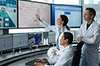 A group of four people, three men and one woman, work together in a control room or monitoring station, with the woman pointing to information displayed on multiple large computer screens. A group of four people, three men and one woman, work together to increase lifecycle efficiency by simulating reality in pharmaceuticals