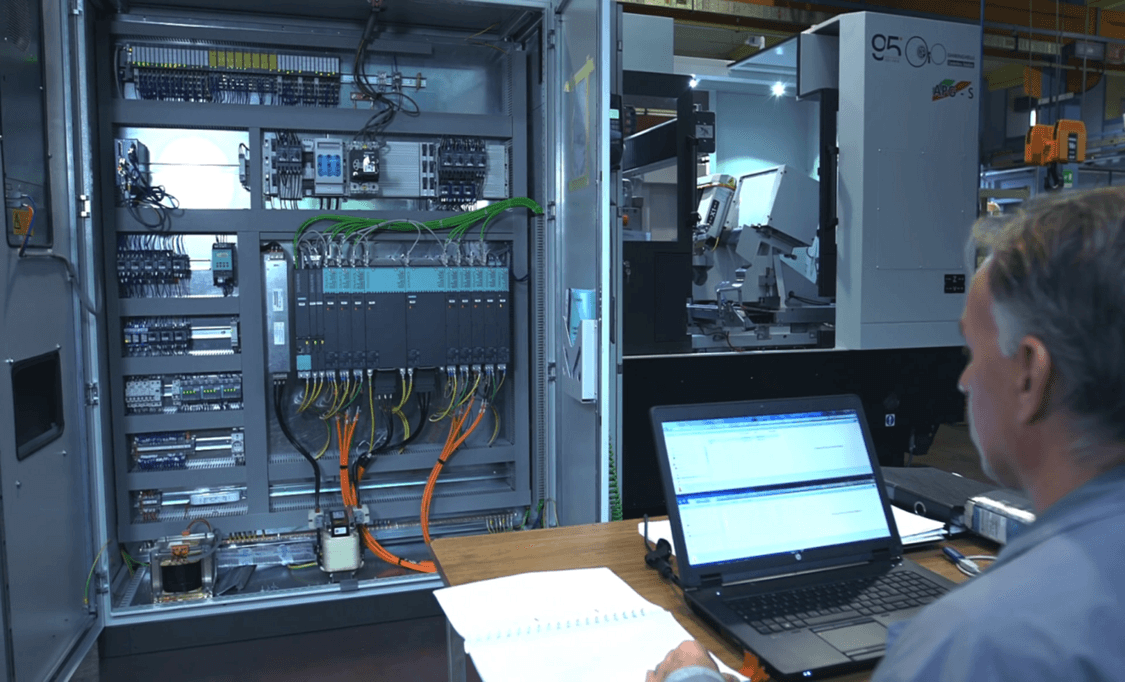 Foto: Inbetriebnahme einer Schleifmaschine, Inbetriebsetzer am Laptop vor offenem, mit Siemens-Komponenten bestücktem Schaltschrank  