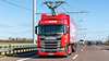 A red truck with current collectors on the roof of the tractor unit on an Autobahn. A catenary system runs overhead.