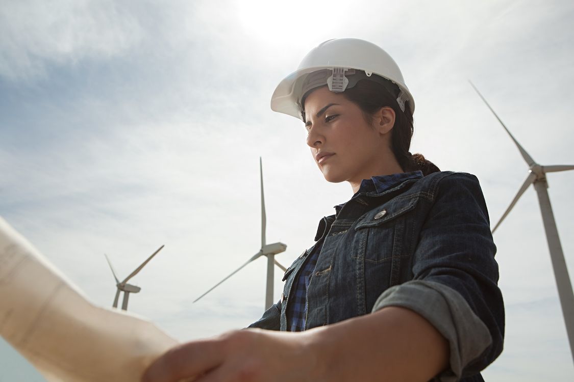 woman engineer wearing hard hat looking at technical drawings with wind turbines in background