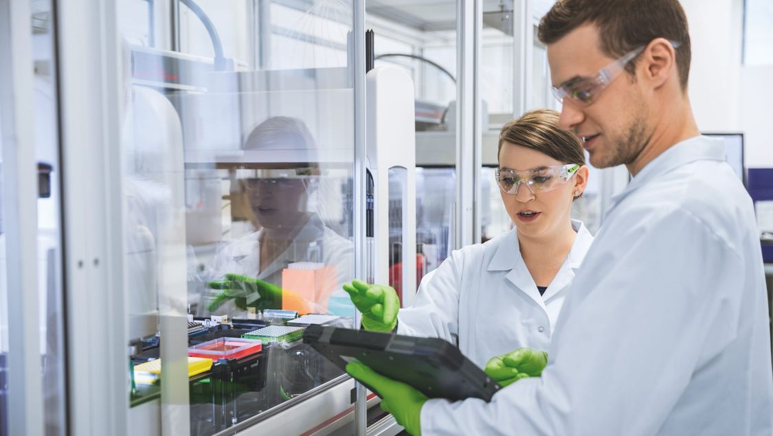 Un hombre y una mujer vestidos de protección trabajan juntos en un Tablet PC industrial en la planta farmacéutica de BioNTech