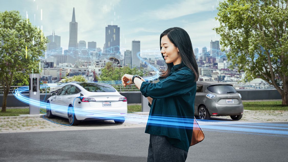  Frau die ein smart device mit ev Autos und Ladegeräten im Hintergrund betrachtet