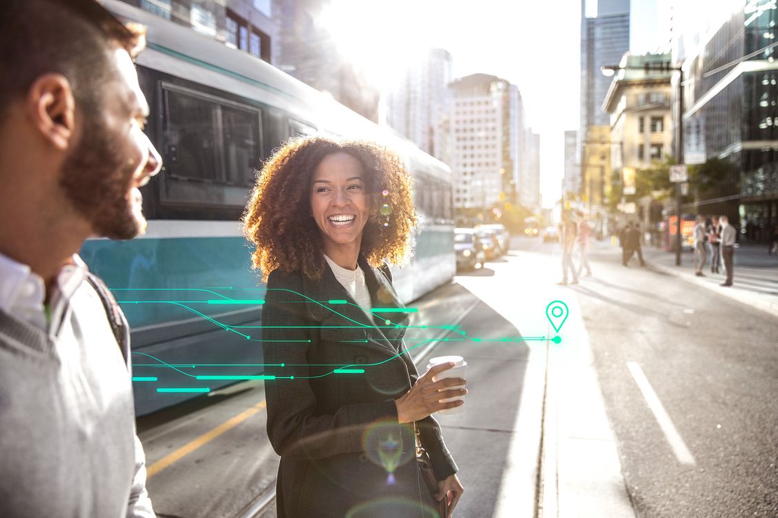 Zwei Personen überqueren lächelnd eine Straße in einer lebendigen, pulsierenden Stadt; hinter ihnen eine Straßenbahn; digitale Elemente über dem Bild zeigen die Möglichkeiten auf, die Smart-City-Lösungen für die Städteplanung bieten.
