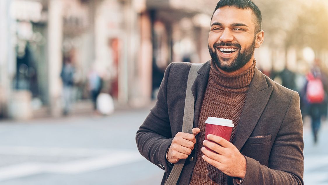 Ein Mann mit Bart Anfang 30 läuft in einer Fußgängerzone. Er trägt einen Kaffeebecher und eine große Umhängetasche.