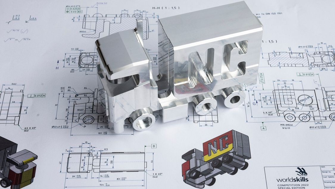 Photo: drawing of a milling workpiece from the competition, placed on it the corresponding real workpiece, a complex truck model.