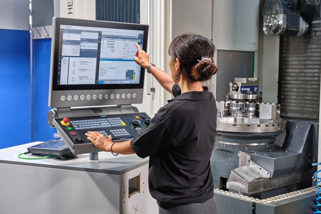 Photo of an employee at a SINUMERIK-One-CNC, in the background the workspace of a 5-axis milling machine with rotary table and clamped complex workpiece