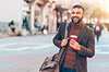 A man with a beard in his early 30s is walking in a pedestrian zone. He holds a coffee cup and is carrying a large shoulder bag. A man with a beard in his early 30s is walking in a pedestrian zone. He holds a coffee cup and is carrying a large shoulder bag.